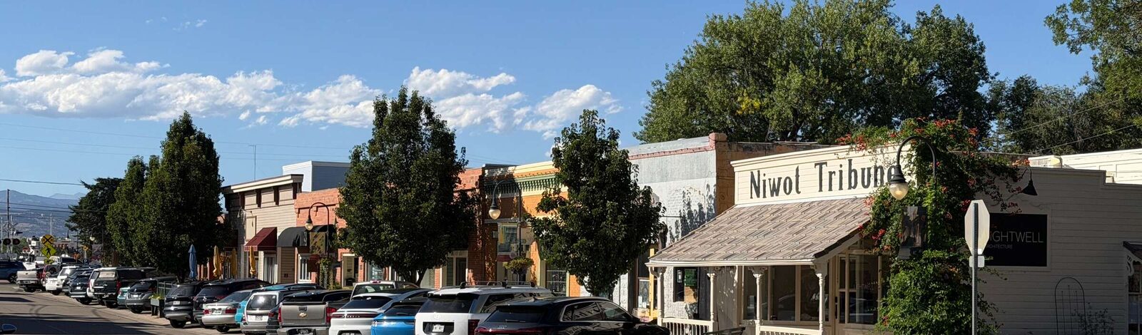 Second Avenue in downtown Niwot on a clear afternoon, looking north along the street. Cars are parked nose-in the whole length of the curb, perhaps two dozen of them. On the right stands the old Niwot Tribune building, white clapboard with its name painted across the front, a shingled porch, a bench outside and a climbing vine in red flower up its corner; beyond it a run of brick and painted storefronts under shade trees, with awnings, a streetlamp and hanging baskets. The Front Range shows low on the horizon at the left.